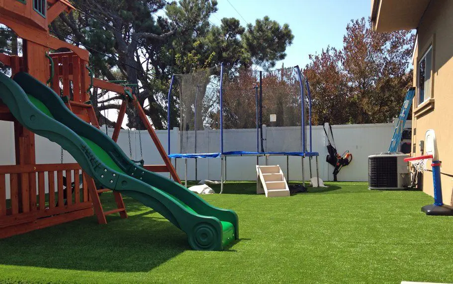 A backyard playground featuring a wooden playset with a slide, a trampoline with safety netting, a basketball hoop, and artificial grass covering the ground. The area is fenced with a white vinyl fence, courtesy of top-notch artificial grass installers from Reno NV, and there's a ladder leaning against the house.