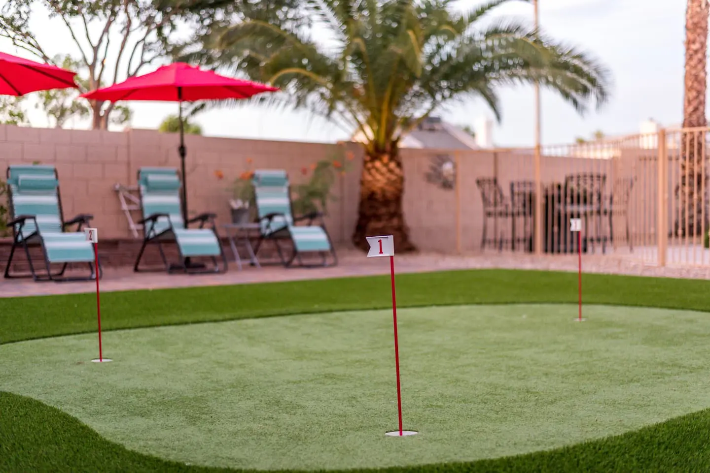 A small backyard putting green with numbered flags on artificial turf, expertly installed by Reno Turf Masters, is surrounded by palm trees and patio furniture. The area includes lounge chairs with umbrellas and a table with chairs, all enclosed by a brick wall and fence.