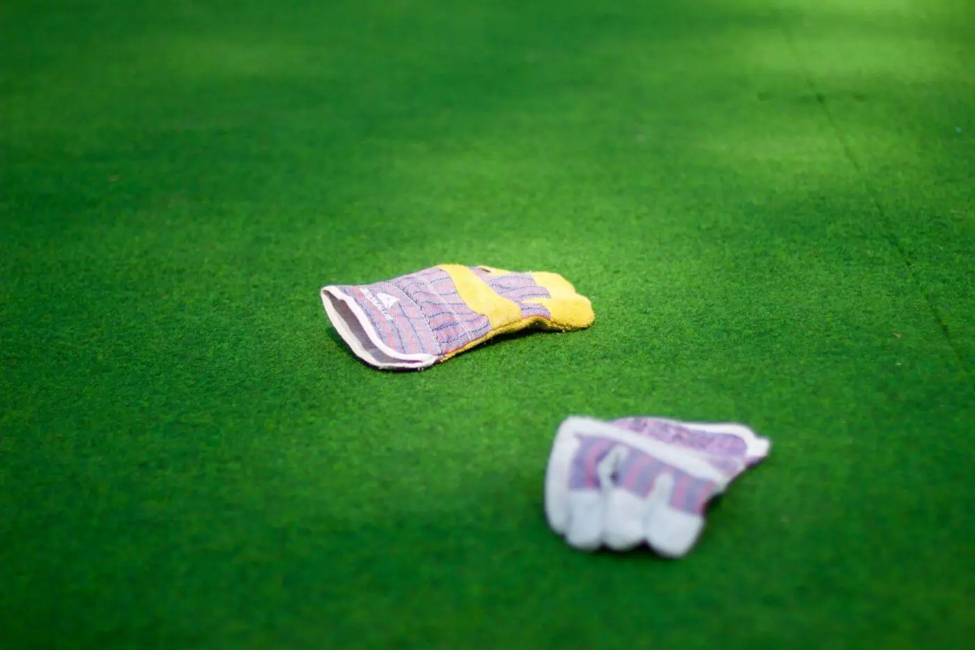 A pair of colorful gardening gloves lies on a well-maintained green lawn, reminiscent of sports fields. The gloves feature a combination of yellow and purple checker fabric. They appear to be casually discarded or left out in the open, possibly by an installation team working nearby.