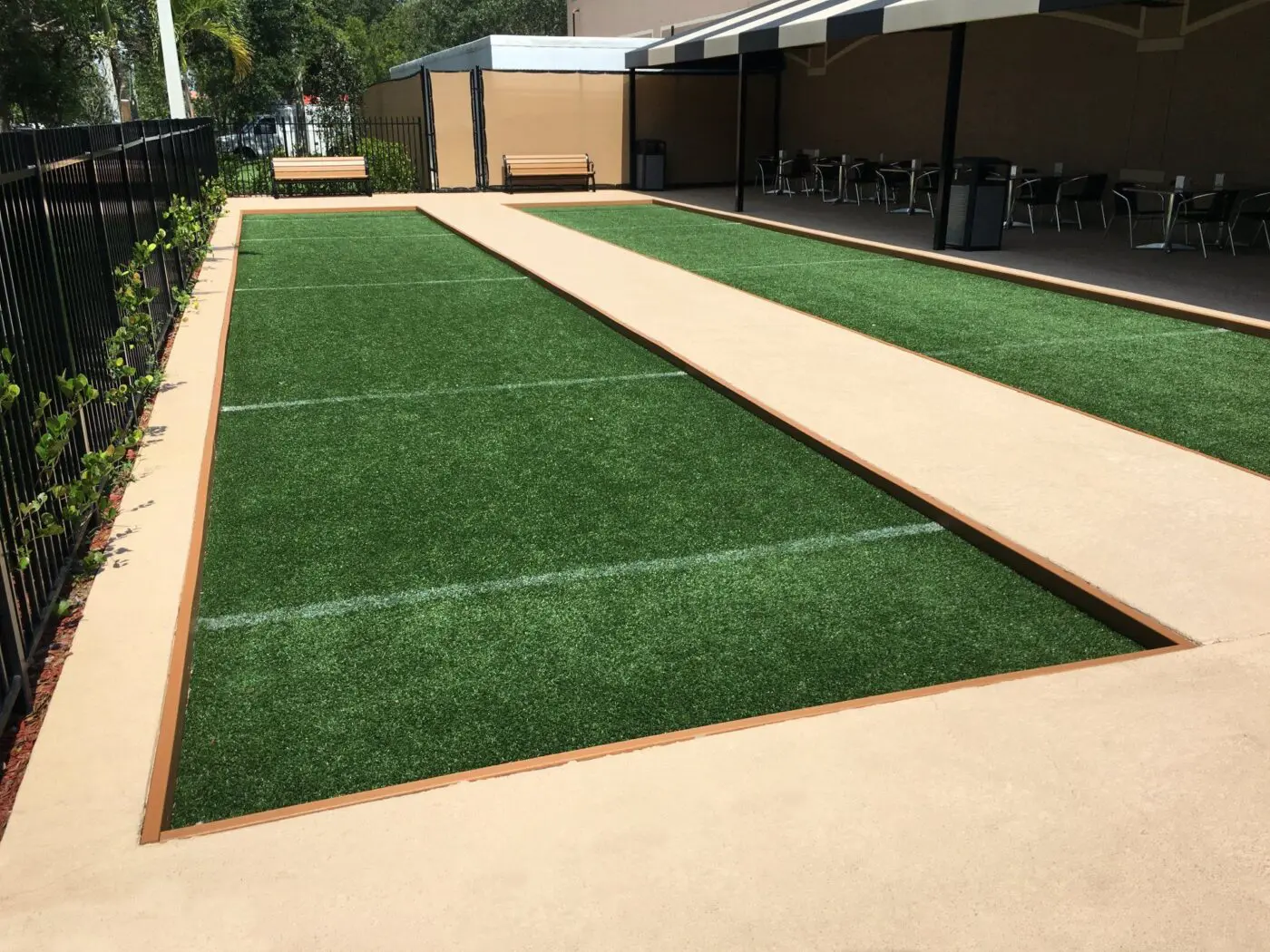 The outdoor bocce ball court features premium artificial turf, bordered by concrete walkways and enclosed by a black metal fence. A wooden bench sits on one side, while tables and chairs rest under a shaded area in the background. The installation team ensured every detail enhances the playing experience.