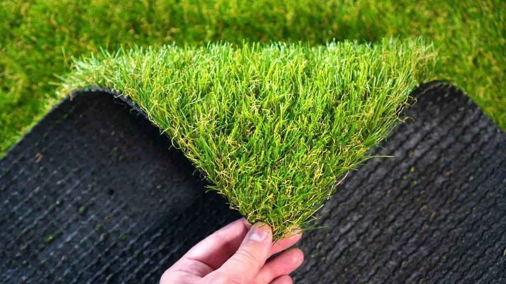 A close-up of a hand lifting a piece of artificial grass, showing the green synthetic turf on top and the black textured backing underneath. The surrounding background also consists of vibrant green grass, perfect for showcasing your Reno NV pet turf installation.