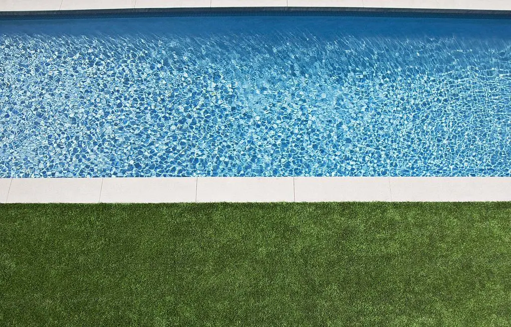 A clear blue swimming pool with rippling water, bordered by a white concrete edge. In the foreground, a lush, green artificial grass lawn provides a backyard surface that requires less maintenance. The image is taken from an aerial perspective, showcasing the pool and the lawn side by side.