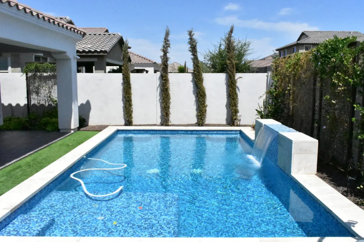 A small rectangular pool with a mosaic tile interior is in a backyard. Water flows from a low, white fountain wall into the pool. The area, featuring artificial grass, is enclosed by a white fence with tall, slender trees. Neighboring houses with tiled roofs are visible in the background. (Reno NV)