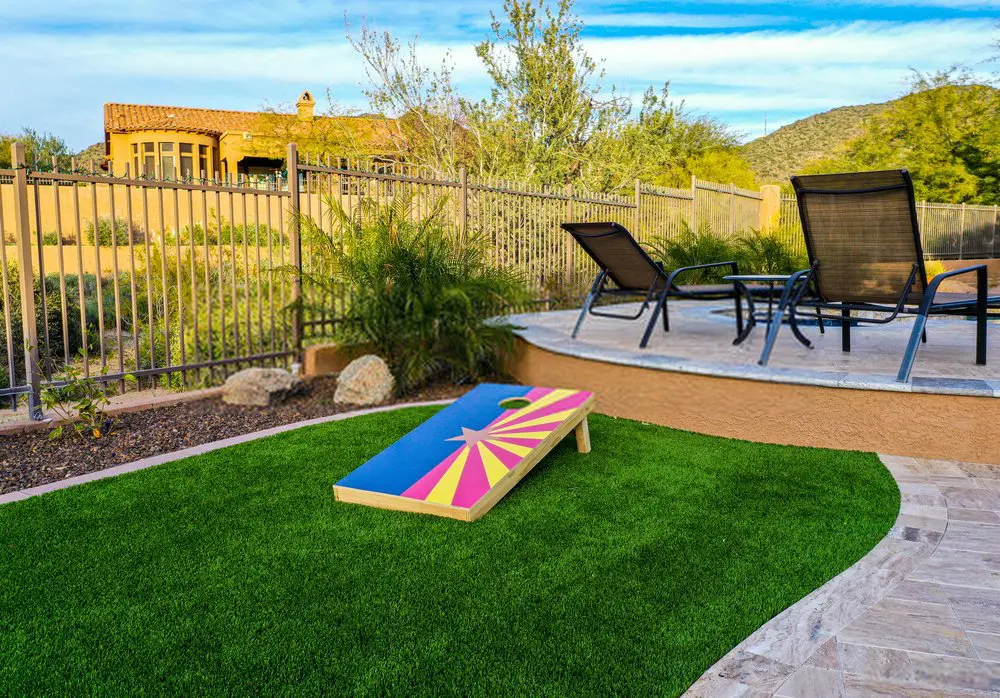 A backyard in Sun Valley NV features a cornhole board with a vibrant design on a patch of artificial grass, installed by an expert artificial grass installer. Lounge chairs rest on the patio enclosed by a fence, with greenery and a house in the background under a blue sky. Free estimates available.