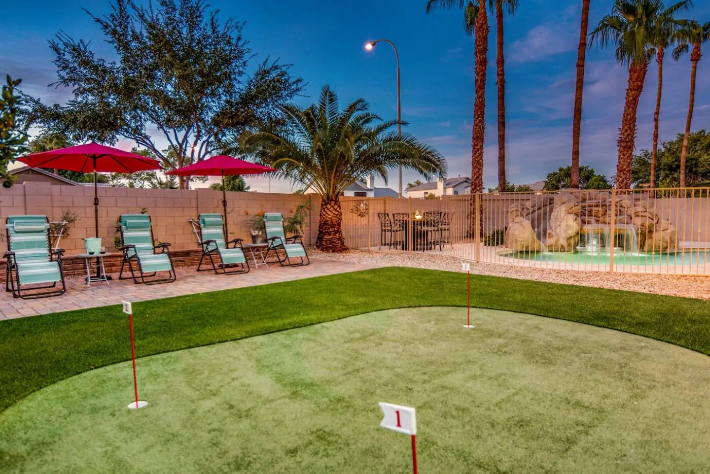 A backyard scene in Sparks NV with a putting green installed by a top-tier artificial grass installer. Three red patio umbrellas over lounge chairs and palm trees enhance the setting. A rock waterfall feature is in the background near a fenced pool area, with evening lighting casting a warm glow.