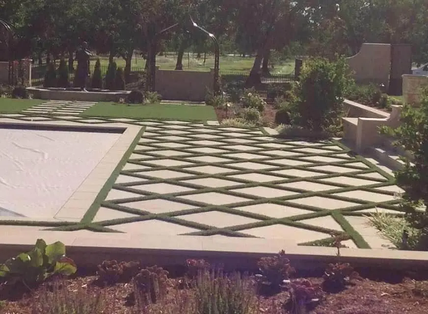 A spacious backyard in Reno, NV, features a geometric pattern of grass strips embedded in a tiled patio, thanks to expert turf installations. A covered outdoor kitchen with stone pillars and an empty rectangular in-ground pool complete the space, which is enclosed by a beige fence and dotted with young trees.