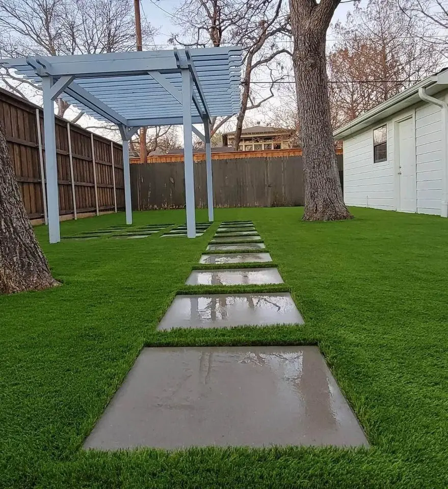 A neatly landscaped backyard with a vibrant green lawn thanks to professional turf installations by Artificial Grass Installers in Reno NV. A row of gray stepping stones creates a path from the house to the wooden fence. Alongside the path, there are small, neatly trimmed bushes and decorative gray pebbles next to a brick wall.