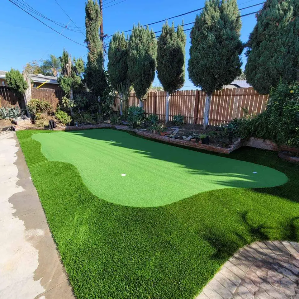 A backyard in Reno NV with a mini putting green featuring two flags, surrounded by a stone patio. In the background, there are stone structures including a raised fire pit and a barbecue grill area against a high fence. Sparse trees and plants are around the border, thanks to professional turf installations by artificial grass installers.