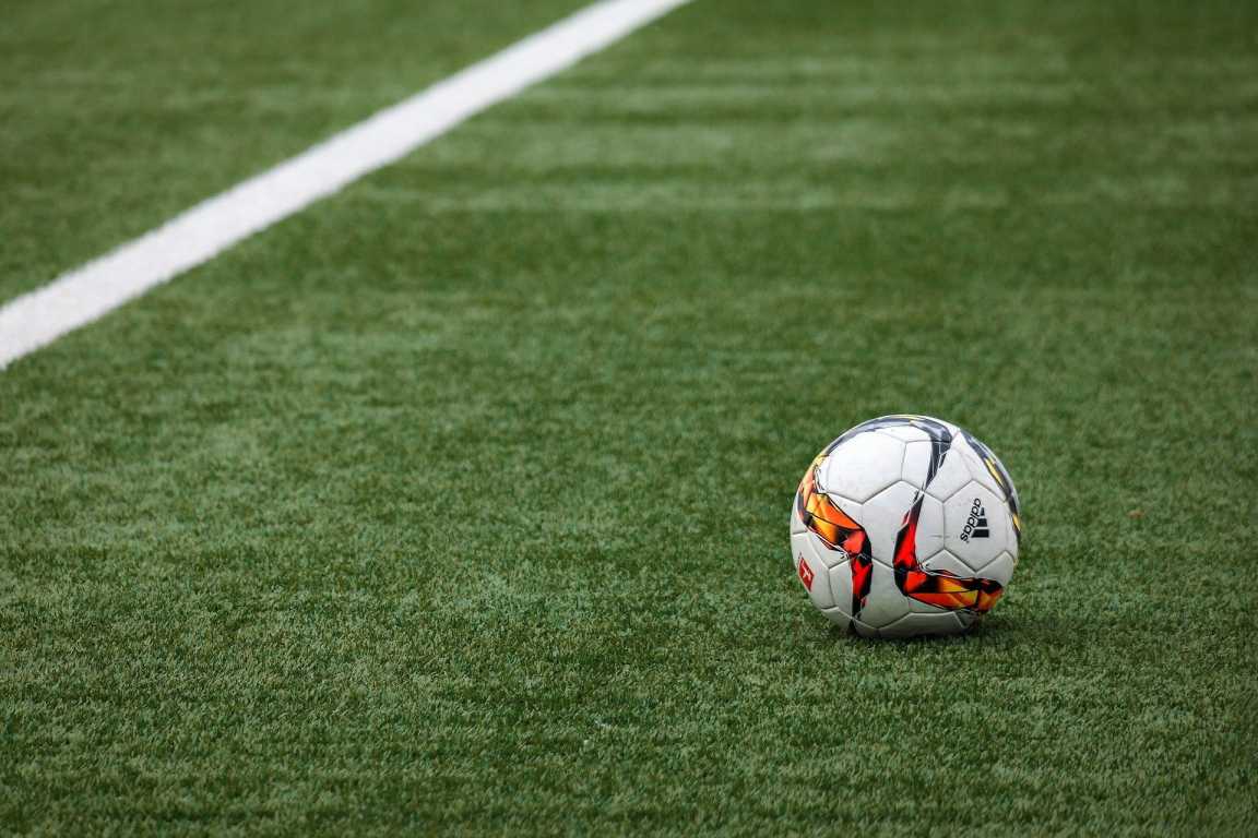 A soccer ball adorned with red, yellow, and black patterns rests on a green, artificial turf field. A white line marking divides the pitch in the background. The scene is set for a game to begin.